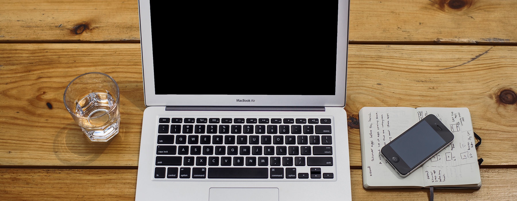 open laptop on desk with glass of water, notebook and cell phone